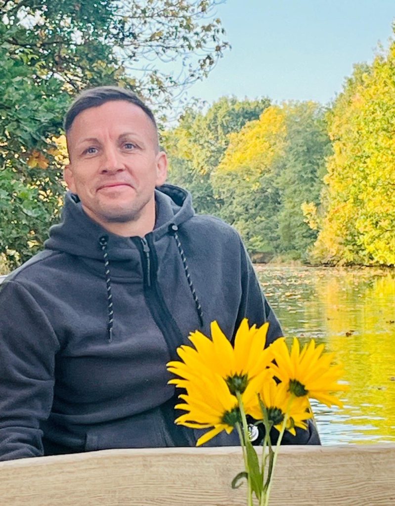 Fährmann Basti lächelnd am Tisch im Kahn, vorn ein Strauß gelber Blumen, dahinter herbstliche Bäume