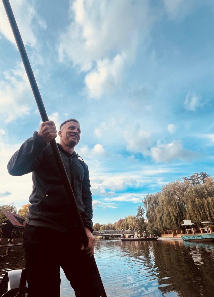 Fährmann Basti stakt den Kahn mit dem Rudel, hinter ihm Trauerweiden und ein Fließ unter blauem Himmel